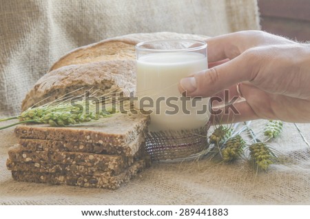 In the foreground is a glass of milk , and in the background bread and wheat spikelets on the burlap. Glass of milk.  Hand taking a glass of milk. Close-up.