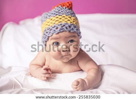 Cool baby, little girl lying on her stomach on the bed, blue eyes, and fun to knit cap on his head
