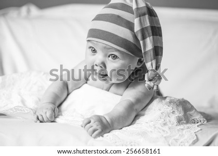 black and white, funny little girl lying on the bed and looking to the side, striped purple stocking on his head, like a gnome, stuck out her tongue, baby