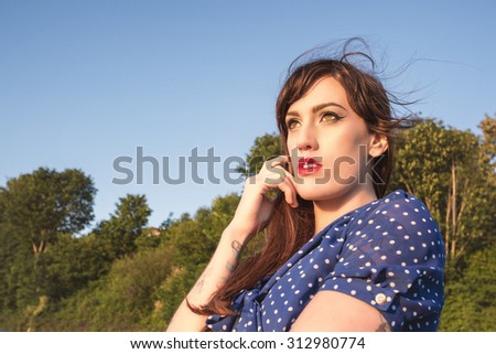 Pretty Young Tattooed Brunette Woman in Blue Polka Dot Dress with Hazel Eyes and Lip Piercing Looking into Distance