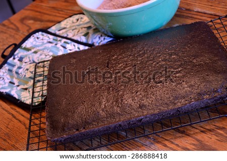 Dark chocolate cake sitting on a cooling rack to cool with colorful oven holders and sea green antique bowl with cake mix in it background on distressed wooden table in old-fashioned kitchen.