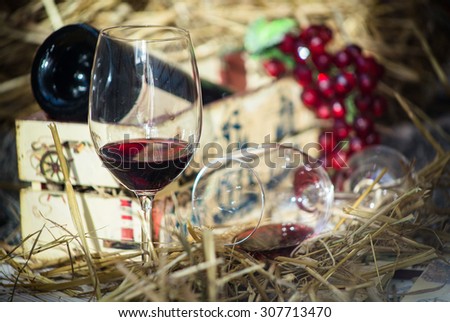 Red wine in a glass, grapes and bottle of wine in a box against a background of hay.