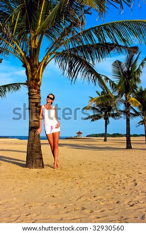 Beautiful brunette girl standing on the lonely sand beach leaning against the palm tree