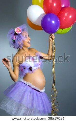 Young beautiful pregnant woman in violet dress with multicolor balloons in studio shot on gray background