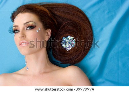 Beautiful red-haired model with long lashes and crystals on face and her hair lying on turquoise silk