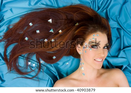 Beautiful red-haired model with long lashes and crystals on face and her long hair lying on turquoise silk