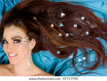 Beautiful red-haired model with long lashes and crystals on face and her long hair lying on turquoise silk