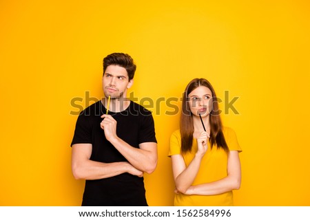 Photo of serious confident couple of two people interested in thinking over standing under empty space holding pens wearing black t-shirt isolated over vivid yellow color background