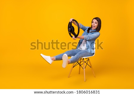 Full body profile photo of pretty funny lady good mood sit chair spread legs playing hold steering wheel riding imagine car wear casual denim shirt shoes isolated yellow color background