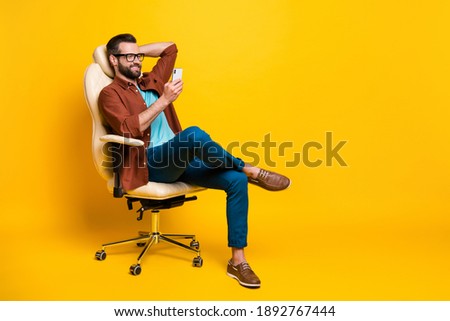 Full length body photo of smiling freelancer keeping smartphone social media sitting in chair pause isolated vibrant yellow color background