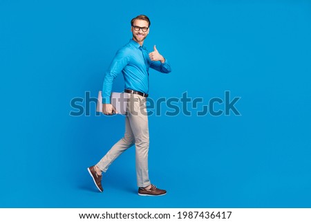 Full size profile side photo of young man happy smile go walk show thumb-up advert advise laptop isolated over blue color background