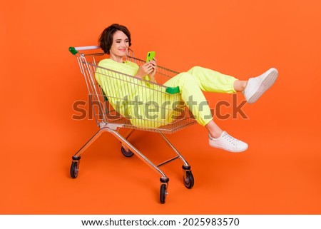 Portrait of attractive cheery funny focused girl sitting in shopping cart chatting blogging isolated over bright orange color background