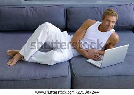 Beautiful young blond man lying down on blue sofa and using laptop.