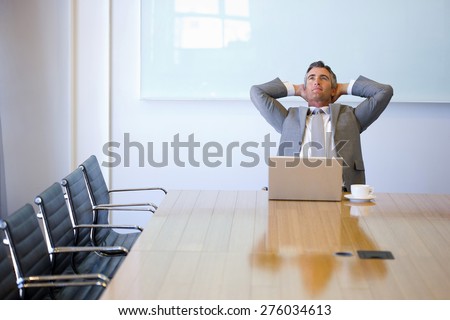 Business manager sitting at the end of a meeting table, with cup of cafe and laptop, relaxing with arms behind head.