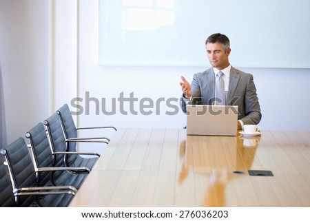 Business manager sitting at the end of a meeting table, with cup of cafe and laptop, moving hands with lecturer attitudes toward empty chairs.