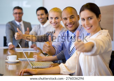 Business team sitting in a line at a meeting table, gesturing thumbs up.