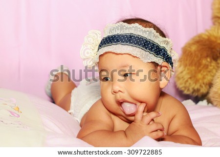 The little baby girl with blue hair band lying prone on the bed pink and hand in she mouth.