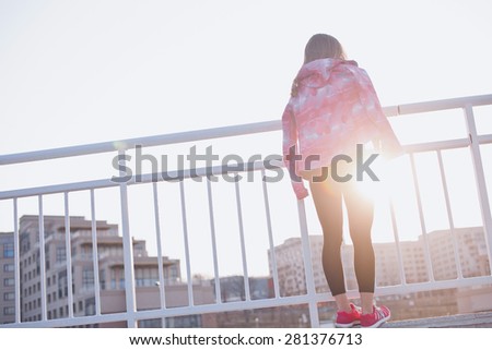 Girl in tights and sportswear at viewpoint stands backside to a camera looking at sunset after training. Urban scenery. Motivation, fitness and healthy lifestyle in a city concept.