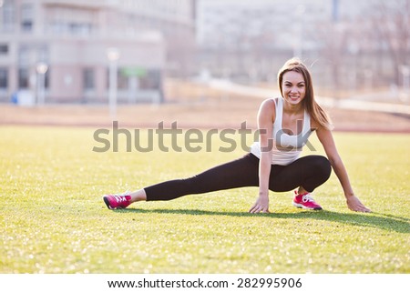 Young woman in sportswear with long hair stretching on a field in a campus. Sit with straight back during exercise. Sunny and bright photo. Concept of healthy lifestyle