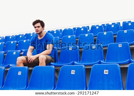 Attractive sporty young man model in blue shirt relaxing on blue stadium seats after training staring at field. Copy space