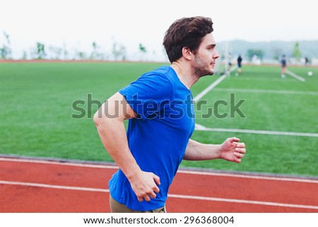 Attractive man runner in blue shirt running fast in front of football field. Success, power and speed concept