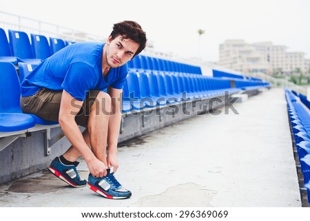 Young attractive male model in blue shirt  tying laces on his running shoes and looking at field before run on a stadium stands after workout perspective view. Copy space