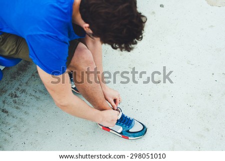 Young attractive male model in blue shirt tying laces on his sports shoes before run on a stadium concrete stands after workout overhead view. Copy space