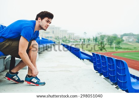 Young attractive male model in blue shirt tying laces on his sports shoes and looking at field before run on a concrete stadium stands after workout. Copy space