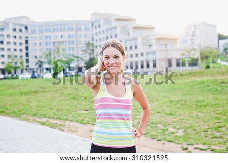 Attractive sporty young woman in striped sleeveless shirt looking at camera smiling and holding her neck while other hand is on belt. After training in university campus park near stadium