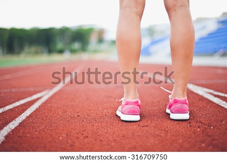 Girl legs in sport shoes standing on a running track with stadium stands and football field on background. Calf cropped back view