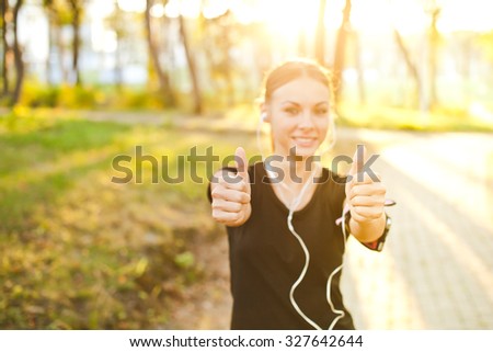 Young attractive sporty female girl with earphones and armband showing thumbs up after successful training on summer evening in park. Sun shining behind model. Copy space. Selective focus on hands