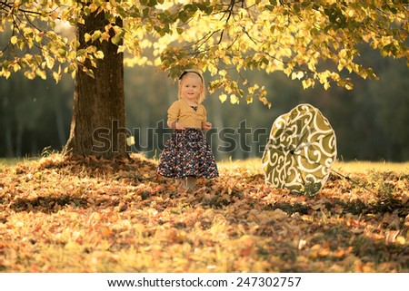 Adorable toddler girl staying under the tree in the park. Green umbrella with white stripes. Autumn. Colorful leaves. Magic back light. Sunset.