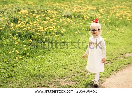 Adorable little girl walking on the dandelion field. White dress with light brown bow. Red ribbon on her head. Big funny glasses on her face. Toned image. Vintage style.