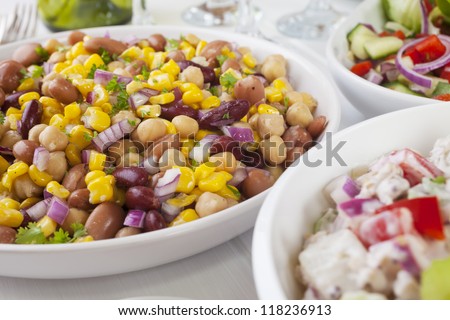 An assortment of salads on a buffet table. Potato salad, bean salad and fresh mixed salad arranged on a white table with glasses, cutlery and plates. Focus on bean salad.
