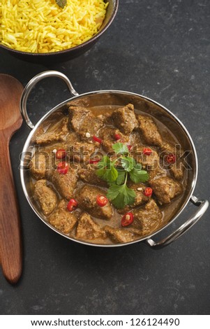 A balti dish or steel karahi with Pork Vindaloo, a hot Indian curry garnished with red chili and coriander, with a side dish of basmati rice.