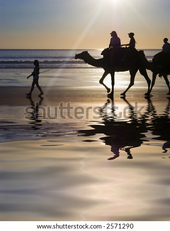 Camel train being led through tidal pools at edge of sea, Cable Beach, Broome, Western Australia. Sun just out of shot causing rays.