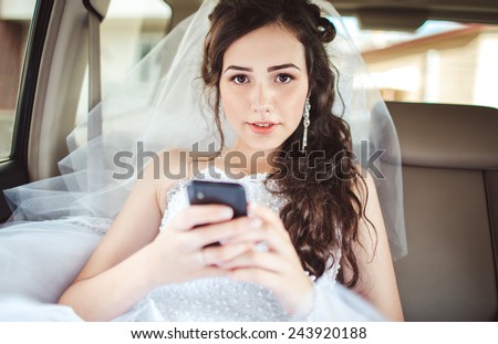 Wedding: beautiful bride sitting in car straight with mobile phone hairstyle and bright makeup. Woman in white dress at wedding day waiting for groom and posing. Concept of love and interest. Newlywed