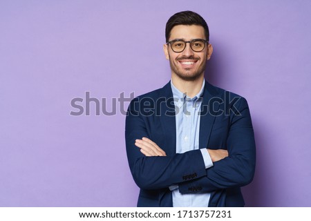 Young buisnessman wearing eyeglasses, jacket and shirt, holding arms crossed, looking at camera with happy confident smile, standing against purple background