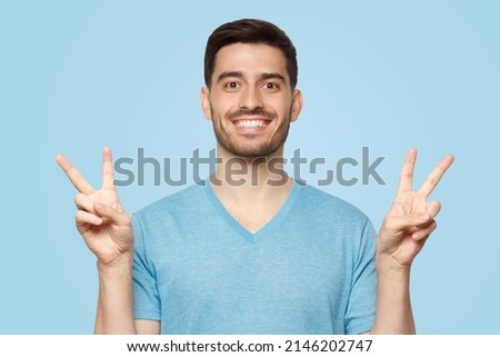 Young man standing in t-shirt, showing victory v sign with fingers of both hands, isolated on blue background  Stock fotó © 