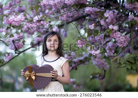 Little brunette smiling girl looking away holding big brown heart shaped present box standing among japanese cherry blossom in the park copyspase, horizontal picture