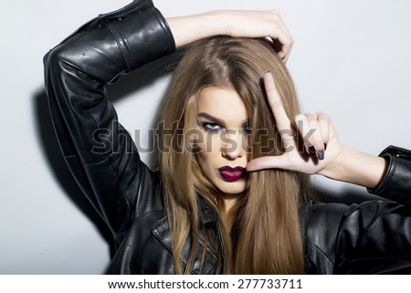 Dangerous sexy young blonde woman with bright make up in black leather jacket looking forward stanging on light grey background closeup copyspace, horizontal picture