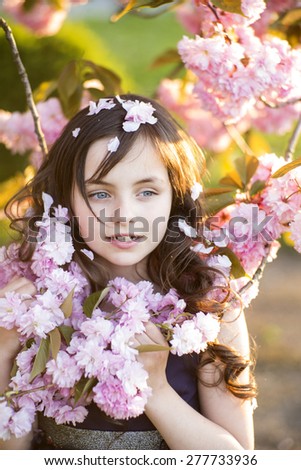 Little brunette girl looking away standing amid pink japanese cherry blossom in broad daylight in the park, vertical picture