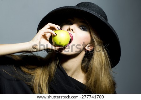 Pretty blonde young woman in retro black hat with bright make up biting fresh green apple standing on gray background copyspace, horizontal picture