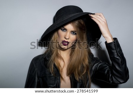 Perilous seductive young blonde girl with bright make up in black leather jacket  and hat looking forward stanging on light grey background closeup copyspace, horizontal picture