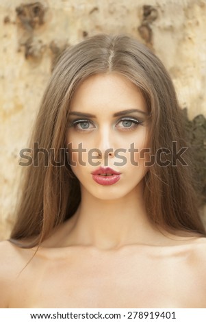 Portrait of pretty young woman with bare shoulders and bright make up looking forward standing in broad daylight outdoor on natural stone background, vertical picture
