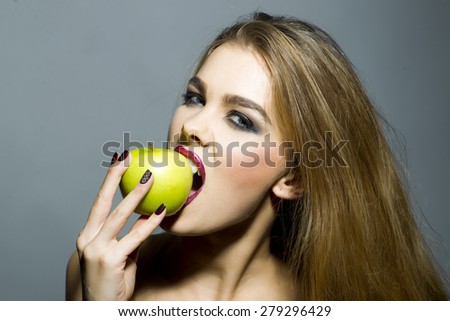 Sensual blonde girl with bright make up looking forward biting fresh green apple standing on gray background copyspace, horizontal picture