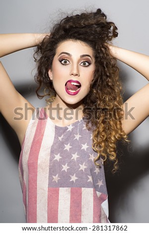 Portrait of happy young crazy girl with curly hair in american flag printed shirt with raised hands standing on light grey wall background, vertical picture
