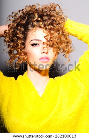 Portrait of fashionable young girl touching her curly hair in yellow sweater looking forward standing on grey wall background, vertical picture