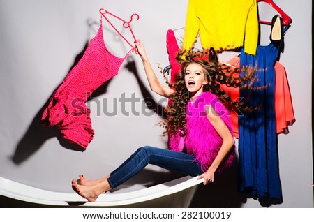 Cool emotional young woman with curly hair in pink fur vest and blue jeans sitting in white bathtub amid colorful clothes pink orange red blue colors on grey wall background, horizontal picture