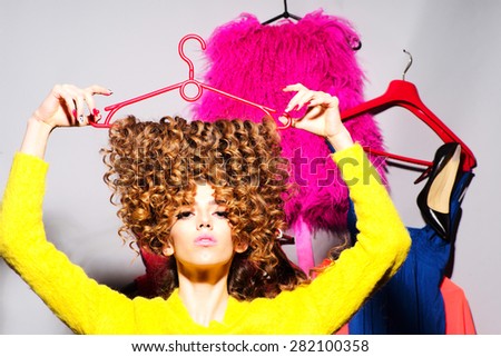 Cool pretty young girl with curly hair in yellow sweater holding hangers standing amid colorful clothes pink red blue colors on grey wall background, horizontal picture